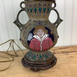 Op de afbeelding: Antieke keramische tafellamp met een gedetailleerd ontwerp. De lamp heeft een bruin en groen kleurenschema met een portret van een persoon in een rode jas. De lamp heeft een houten voet en decoratieve accenten.