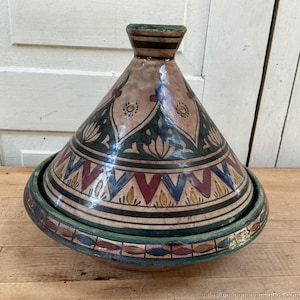 May include: A ceramic tagine pot with a conical lid and a wide, shallow base. The pot features a beige background with intricate patterns in dark green, red, blue, and yellow. The design includes geometric shapes and floral motifs, creating a decorative and functional kitchenware item.
