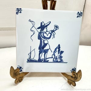 May include: A square white ceramic tile featuring a blue hand-painted illustration of a person wearing a wide-brimmed hat and blowing into a horn. The design includes small floral corner accents, a sailboat, and a bush in a blue ink style.