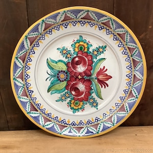 May include: A large, white ceramic plate with a yellow rim and a colourful floral design. The plate features a border of blue, purple and brown triangles and a repeating pattern of yellow and blue lines.
