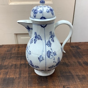 Puede incluir: Cafetera de porcelana blanca con tapa y asa. La jarra está decorada con motivos florales azules y detalles acanalados verticales. El pico está en el lado izquierdo y el asa es curvada. La jarra se apoya sobre una base blanca.