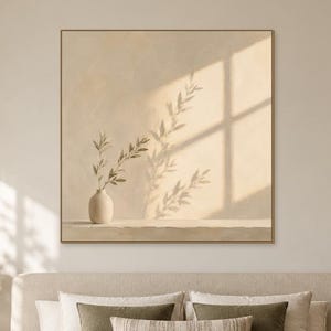 May include: A square framed wall art print featuring a beige background with a small vase containing a leafy branch and its elongated shadow. The artwork is displayed above a bed with beige bedding, olive green pillows, and a matching throw blanket.