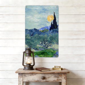 May include: Metal sign displaying a reproduction of Van Gogh's "Starry Night" painting. The artwork shows a vivid night scene with a bright yellow moon, dark cypress trees, and rolling hills. The sign is above a wooden table with a lantern, book, and brass object.