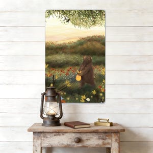 May include: A rectangular metal sign depicting a brown bear with a glowing yellow object in a wildflower meadow. The background shows a sunset and a tree branch with a bird. The sign is displayed on a wooden table.