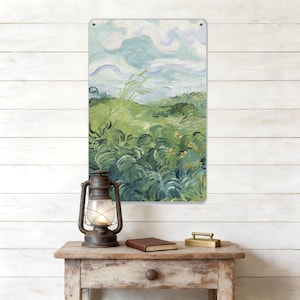 May include: A metal sign displaying a landscape painting with green fields, a blue sky, and white clouds. The sign is mounted on a white wooden wall. A vintage lantern, a brown book, and a brass paperweight are on a wooden table.