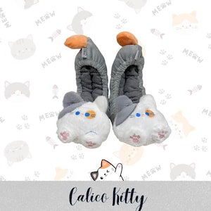 May include: A pair of cat-themed slippers. The slippers are white with gray accents and feature embroidered cat faces with blue eyes and orange ears. The slippers have a gray band that goes over the foot and an orange pom-pom on top. The text "Calico Kitty" is at the bottom.