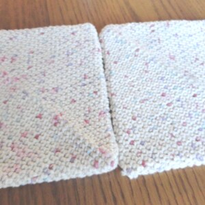 May include: Two off-white, square knitted washcloths with a speckled pattern of pink, purple, and blue. The washcloths are side-by-side on a wooden surface. The knitted texture is visible.