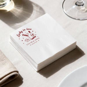 Op de afbeelding: Een stapel witte papieren servetten met een bordeauxrood ontwerp. Het ontwerp bevat de tekst "Lucky in love" en illustraties van een martini, fles, speelkaarten en harten. De namen "LUCAS & ANNA" en een datum staan ook afgedrukt.