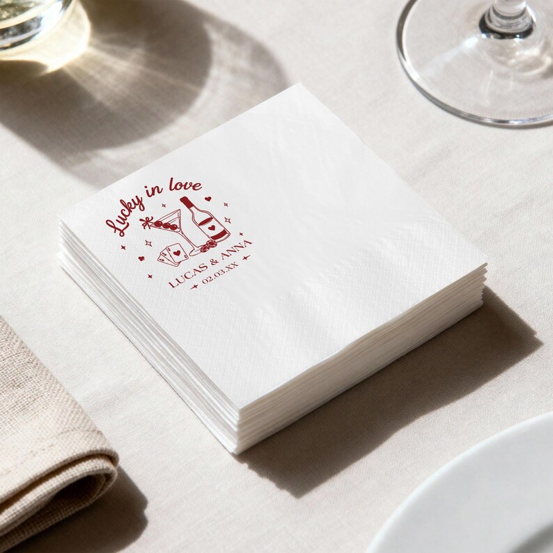 Op de afbeelding: Een stapel witte papieren servetten met een bordeauxrood ontwerp. Het ontwerp bevat de tekst "Lucky in love" en illustraties van een martini, fles, speelkaarten en harten. De namen "LUCAS & ANNA" en een datum staan ook afgedrukt.