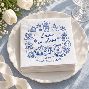 Op de afbeelding: Een vierkant wit papieren servet met blauwe inktillustraties van wintersportuitrusting, een sneeuwpop, een kampvuur en bergen. De tekst luidt Snow in Love, est 2026 en Sophie's Bach | Vail, CO, gerangschikt in een cirkelvormig patroon.