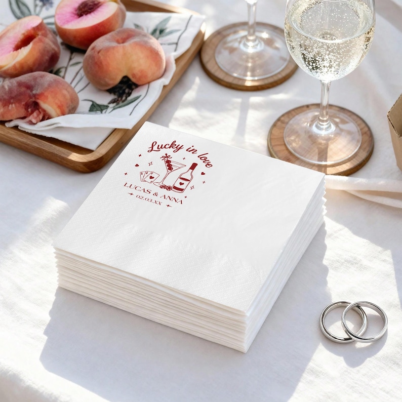 Op de afbeelding: Een stapel witte papieren servetten met een rode afbeelding en de tekst "Lucky in love" met een cocktail, speelkaarten en een flesillustratie. Twee zilveren trouwringen staan op de voorgrond. Perziken en champagneglazen staan op de achtergrond.