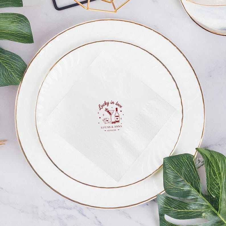 Op de afbeelding: Witte keramische borden met gouden randen, de ene op de andere gestapeld, met een witte servet met de tekst "Lucky in love" en een cocktailillustratie. Groene bladeren staan op de voorgrond.