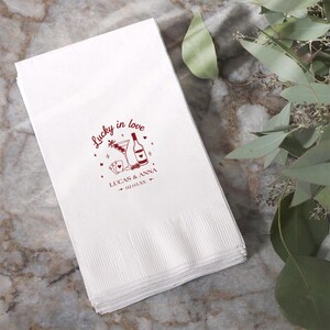 Op de afbeelding: Witte papieren servetten met een rood ontwerp met de tekst "Lucky in love", een martiniglas, een fles, speelkaarten, de namen "Lucas & Anna" en een datum. De servetten zijn opgestapeld en hebben een textuur.