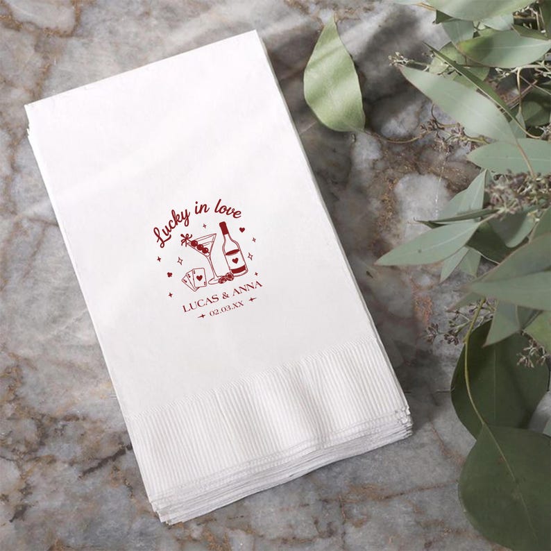 Op de afbeelding: Witte papieren servetten met een rood ontwerp met de tekst "Lucky in love", een martiniglas, een fles, speelkaarten, de namen "Lucas & Anna" en een datum. De servetten zijn opgestapeld en hebben een textuur.