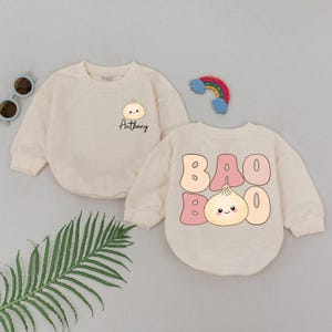 May include: Two cream-colored baby outfits. One features a dumpling graphic with the name "Anthony" in black script. The other has the words "BAO BOO" in pink bubble letters with a dumpling graphic. A rainbow and sunglasses are in the background.