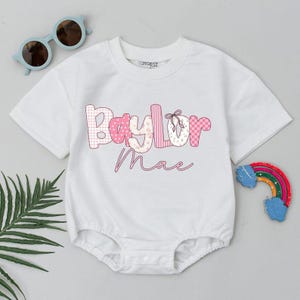 Op de afbeelding: Witte babybody met de naam "Baylor Mae" in roze en witte patchwork letters. Het ontwerp bevat een ballet-schoenafbeelding. Lichtblauwe zonnebrillen en een regenboogdecoratie zijn ook zichtbaar.