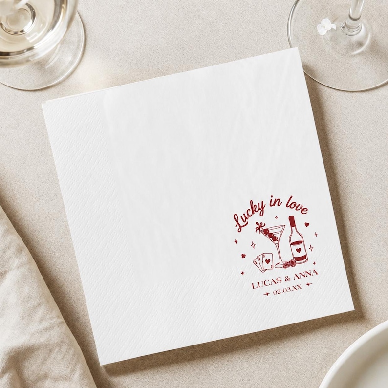 Op de afbeelding: Witte papieren servet met een getextureerd oppervlak. De servet heeft een bordeauxrode afbeelding met de tekst "Lucky in love" en illustraties van een martini, speelkaarten en een fles wijn. De namen "Lucas & Anna" en een datum zijn ook opgenomen.