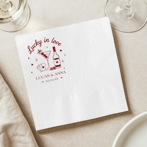 Op de afbeelding: Witte vierkante papieren servet met een rood grafisch ontwerp. Het ontwerp bevat een martiniglas, een wijnfles, speelkaarten en de tekst "Lucky in love" en "Lucas & Anna 02.03.XX".
