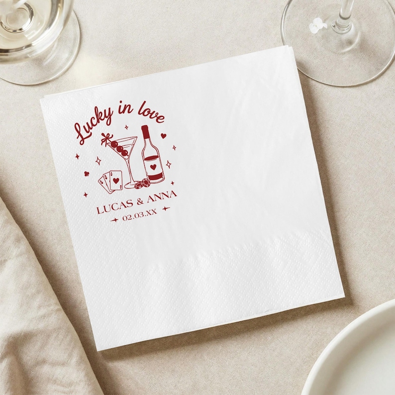 Op de afbeelding: Witte vierkante papieren servet met een rood grafisch ontwerp. Het ontwerp bevat een martiniglas, een wijnfles, speelkaarten en de tekst "Lucky in love" en "Lucas & Anna 02.03.XX".