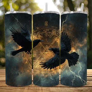 May include: Three stainless steel tumblers with black raven designs against a dark, stormy background. The ravens are in flight, with a golden, circular design in the center tumbler. Each tumbler has a clear lid and a straw.