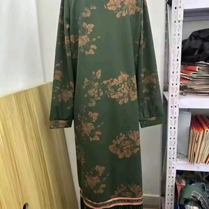 以下が含まれることがあります： オリーブグリーンの長袖膝丈チュニック。銅色の花柄が全体に施されています。襟元には装飾的な縁取りがあり、裾の上部には同色のトリムがあしらわれ、一番下には長い黒いフリンジが付いています。