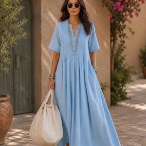 Könnte beinhalten: Ein hellblaues Maxikleid mit kurzen Ärmeln, V-Ausschnitt und vertikalen Falten am Oberteil. Das Kleid hat einen locker fallenden Rock und wird mit einer großen, cremeweißen Canvas-Tasche, geschichteten Goldketten und braunen Sandalen kombiniert.