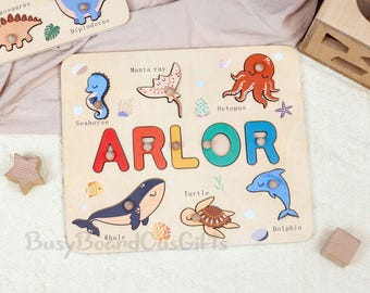 May include: Wooden puzzle featuring the name "ARLOR" in colorful letters, with sea creature cutouts like a seahorse, whale, and dolphin. The puzzle is designed for children and includes a wooden star and block.