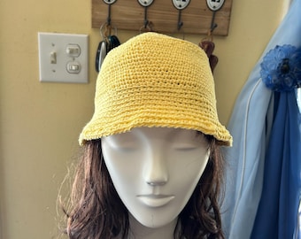 May include: A yellow crocheted bucket hat. The hat is made of a textured yarn and has a slightly curved brim. The hat is displayed on a mannequin head with brown hair. The background includes a wall with a key holder and a light switch.
