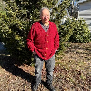 May include: A red, button-down cardigan sweater with a textured knit pattern. The sweater has wooden buttons and is worn with a gray shirt and black jeans. The person is standing outside in front of a green tree.
