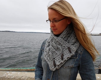 May include: A chunky grey crochet cowl scarf featuring a cable knit pattern and three dark brown wooden toggle buttons arranged vertically along the front edge, worn over a blue denim jacket with visible stitching and metal button details.