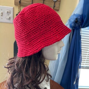 May include: A bright red, handmade crochet bucket hat. The hat is displayed on a mannequin head with brown hair. The hat has a slightly flared brim and a close-knit texture, showcasing the detailed crochet work.