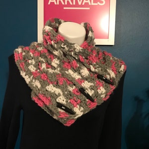 May include: A handmade crochet cowl in gray, white, and pink, fastened with three black toggle buttons. The cowl is displayed on a mannequin, with a sign in the background that reads "NEW ARRIVALS".