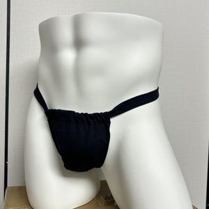 Puede incluir: Tanga negro sobre un maniquí blanco. La ropa interior tiene una bolsa delantera y correas finas que se atan alrededor de la cintura. El maniquí es una forma de torso, sin brazos ni piernas.
