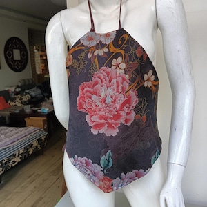 Könnte beinhalten: Ein Neckholder-Top mit floralem Design. Das Top hat eine große rosa Pfingstrose in der Mitte, umgeben von kleineren Blüten in Rosa, Weiß und Blau. Der Hintergrund ist dunkel und gedämpft. Die Träger sind dunkelbraun.