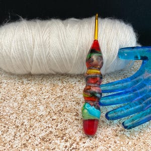 May include: A colorful glass crochet hook with a red, yellow, blue, and green swirl pattern. The hook is resting on a white ball of yarn and a blue plastic yarn bowl.