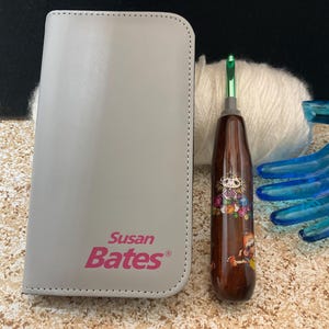 May include: A light gray Susan Bates crochet hook case and a wooden crochet hook with a colorful design. A ball of white yarn and a blue decorative hand are also in the image.