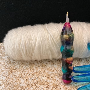 May include: A crochet hook with a thick, ergonomic handle featuring a swirling, multi-colored resin pattern in shades of pink, teal, black, and gold. The hook has a metallic tip and is positioned vertically in front of a ball of white yarn.
