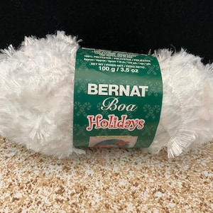 May include: A skein of white Bernat Boa Holidays yarn with a fuzzy, feathery texture. The green paper label features the brand name, product name, and states 100% polyester and a net weight of 100 g / 3.5 oz. The yarn has a soft, shaggy appearance.