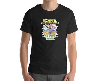 Può includere: T-shirt grigio scuro con un mostro dei cartoni animati a una scrivania con la scritta "I Can't Tax Season Is Over". Il mostro è viola con corna gialle ed è circondato da fogli di carta. Il testo è giallo e bianco.