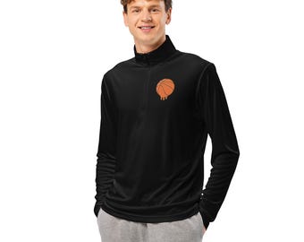 Può includere: Maglia sportiva nera a maniche lunghe con colletto con zip. La maglia presenta una grafica di pallone da basket arancione con dettagli gocciolanti sul petto. Il modello indossa pantaloni della tuta grigi.