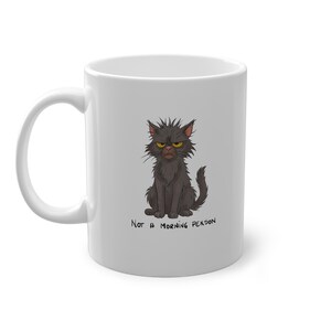 Könnte beinhalten: Weiße Keramiktasse mit einer Illustration einer grimmigen schwarzen Katze mit gelben Augen. Der Text "Not a Morning Person" steht unter der Katze. Die Tasse hat einen gebogenen Griff.