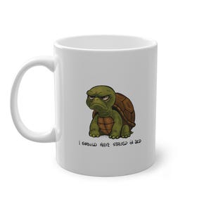 May include: White ceramic mug featuring a grumpy cartoon turtle with a brown shell and green body. The text "I SHOULD HAVE STAYED IN BED" is printed below the turtle. The mug has a curved handle.