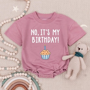 May include: Dusty rose-colored baby romper with the text "NO, IT'S MY BIRTHDAY!" and a cupcake graphic with a lit candle. The short-sleeved one-piece has snap closures and a round neckline. A teddy bear and decorative beads are in the background.