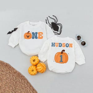 May include: White long-sleeved baby rompers with pumpkin graphics. One romper has the word "ONE" and a pumpkin graphic. The other has the name "HUDSON" and a pumpkin with the number "1". Halloween-themed baby clothing.