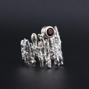 May include: A silver-colored ring with a textured, organic design. The ring features a small, round, deep red gemstone set in a bezel. The ring's surface has a rough, uneven texture, giving it a unique, artistic appearance.