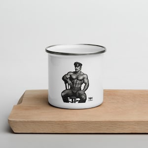 Può includere: Tazza smaltata bianca con un'illustrazione in bianco e nero di un uomo muscoloso con un berretto seduto su una sedia. La tazza ha un bordo argentato e un manico. L'immagine è su un tagliere di legno.