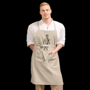Op de afbeelding: Een beige schort met een tekening van een gespierde man. De schort heeft een strik in de taille en twee voorzakken. De persoon op de foto draagt een wit overhemd.