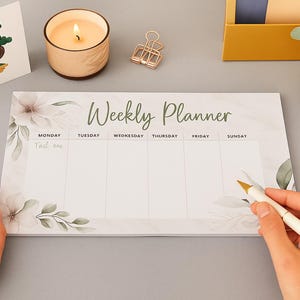 May include: A weekly planner with floral designs and the words "Weekly Planner" in green script. A lit candle, pens, and a decorative card with a bonsai tree are also visible. A hand is holding a pen.