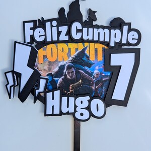 Op de afbeelding: Een zwart-witte Fortnite-thema taarttopper met de tekst "Feliz Cumple Hugo 7". De topper heeft een kleurrijke afbeelding van personages uit het spel en bliksemaccenten. Een zwarte stok is bevestigd om in een taart te steken.