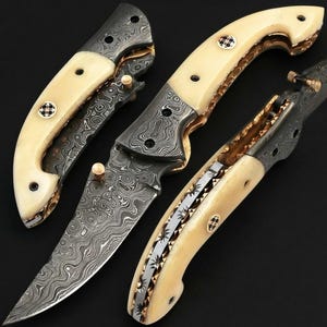 Könnte beinhalten: Drei Klappmesser mit Damaststahlklingen mit ausgeprägtem Wellenmuster. Die Griffe bestehen aus glattem, cremefarbenem Material, akzentuiert durch dekorative Messingstifte und aufwendige Metallfeilarbeiten entlang des Griffrückens.